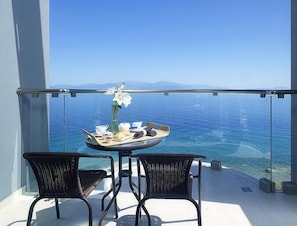 Een balkon met een ontbijttafel, twee rieten stoelen en een helder uitzicht op de zee en verre bergen.