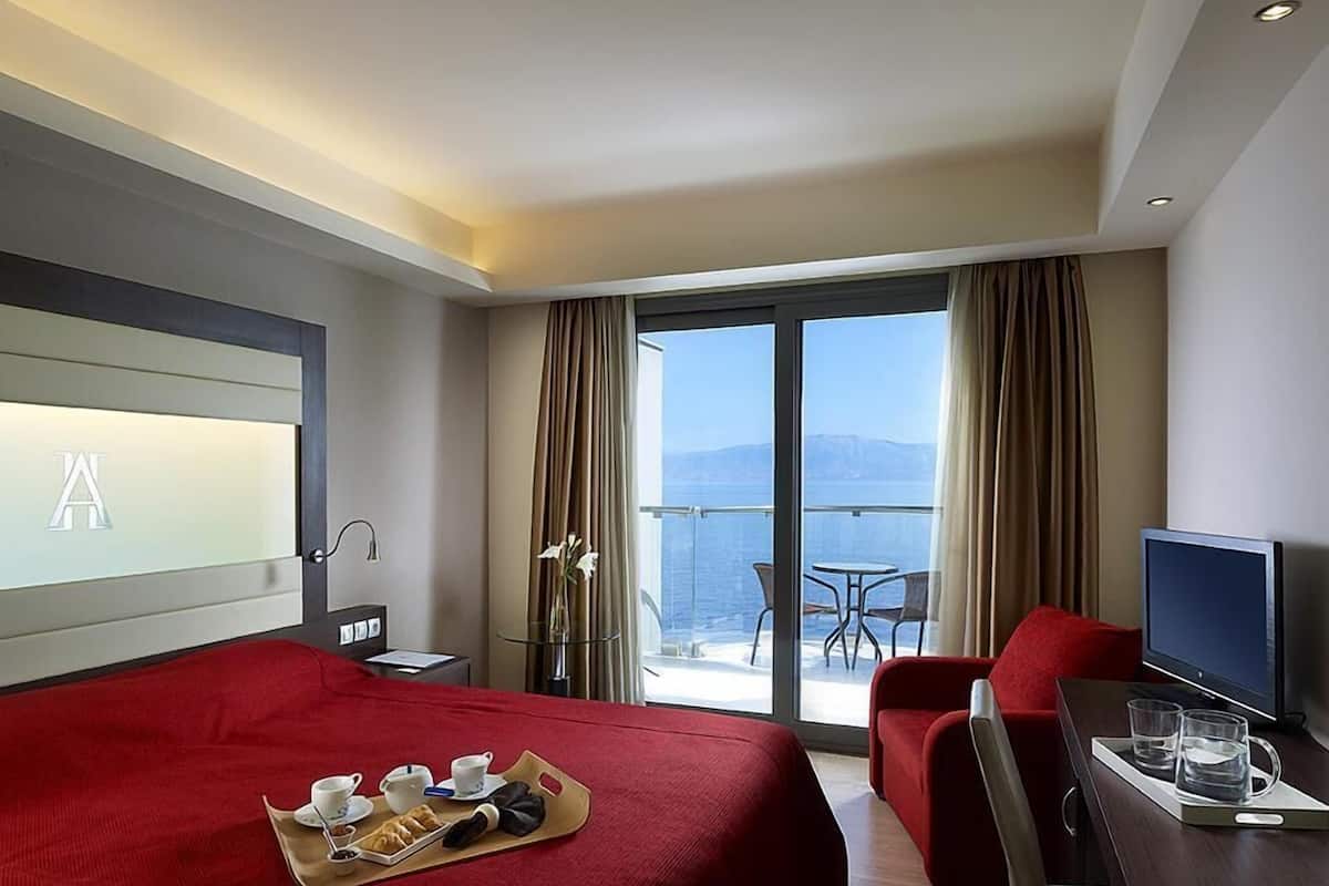 Een hotelkamer met een bed, een bureau met een televisie, een balkon met een tafel en stoelen, en uitzicht op de zee.