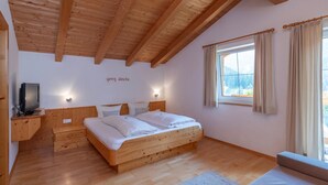 A bedroom with a wooden ceiling, a bed with white bedding, a television on a wooden stand, and a window with curtains.