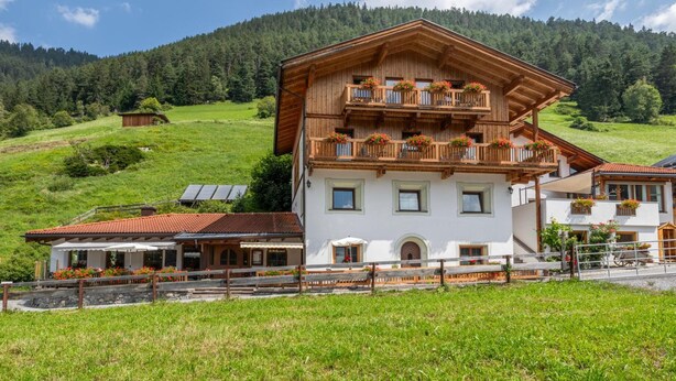 A two-story chalet with a balcony, surrounded by a lush green landscape and a forested mountain in the background.