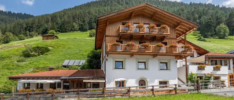 A two-story chalet with a balcony, surrounded by a lush green landscape and a forested mountain in the background.