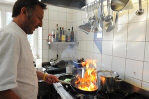 A chef cooking with a pan on a stove with flames, kitchen utensils hanging above, and bottles of alcohol on a shelf.