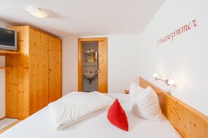 A bedroom with a wooden wardrobe, a bed with white and red pillows, a wall-mounted light fixture, and a television on the left.