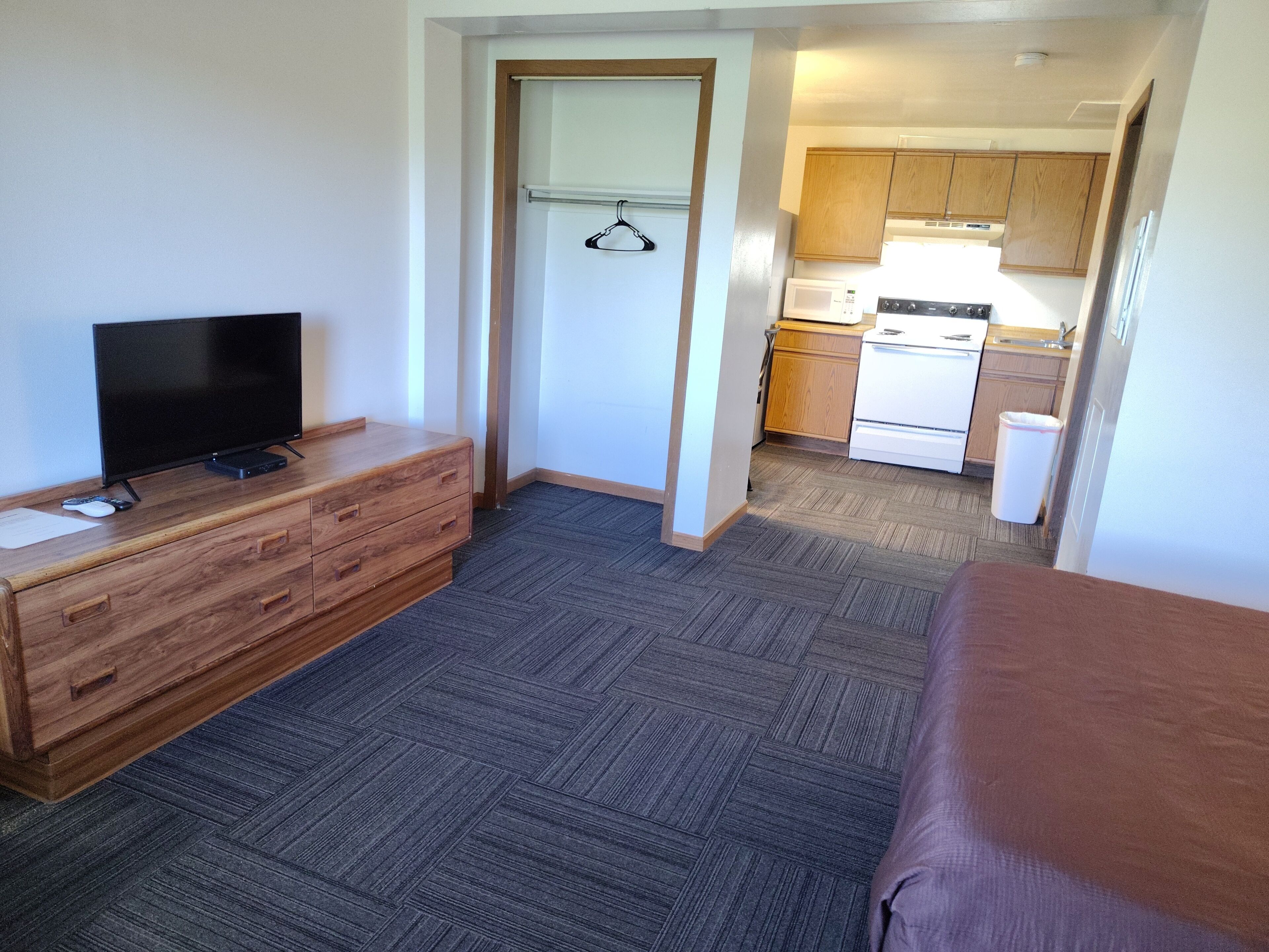 A compact hotel room with a television, wooden dresser, a bed, a kitchenette, and a door leading to a bathroom.