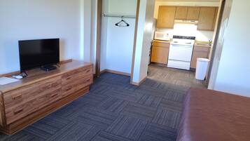 A compact hotel room with a television, wooden dresser, a bed, a kitchenette, and a door leading to a bathroom.