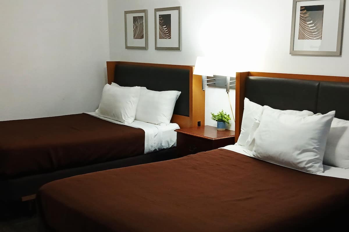 A hotel room with two single beds, a nightstand with a lamp, and framed artwork on the wall.