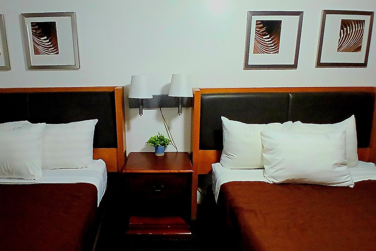Habitación de hotel con dos camas, una mesita de noche con lámpara y cuadros en la pared.