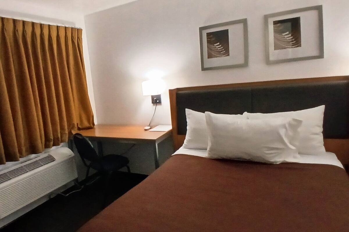 Una habitación de hotel con cama, escritorio, silla, lámpara y aire acondicionado instalado en la pared.