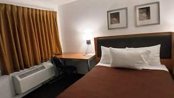 Una habitación de hotel con cama, escritorio, silla, lámpara y aire acondicionado instalado en la pared.