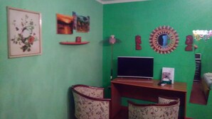 Una habitación con pared verde, un escritorio de madera con una computadora, dos sillas con diseño y varios adornos en la pared, incluyendo un espejo, un cuadro enmarcado y una estantería con objetos.