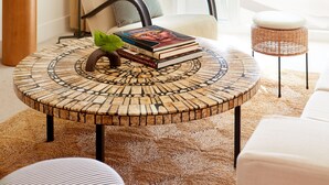 A modern living room with a round coffee table, a white sofa, and a chair. There is a large window with sheer curtains, a potted plant, and a view of the ocean. - Palm Heights (Seven Mile Beach)