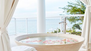 A bathtub with floral bath products, a wooden stool, and a pair of slippers on a balcony with a view of the sea. - Palm Heights (Seven Mile Beach)