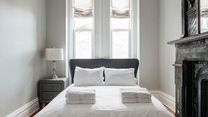 Un lit bien fait, avec du linge de lit blanc et deux oreillers, une table de chevet avec une lampe, et une cheminée surmontée d’un manteau sombre et orné.