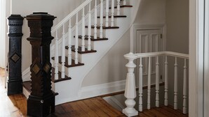 Un escalier en bois, doté de balustrades et d’une main courante blanches, mène à un palier où se trouve une colonne en bois foncé et un luminaire suspendu.