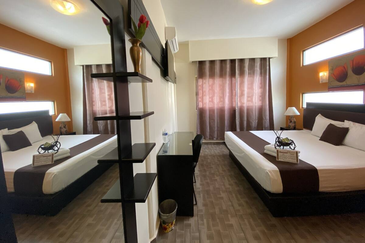 A hotel room with two beds, a small table, a vase with flowers, a TV, and a window with curtains.