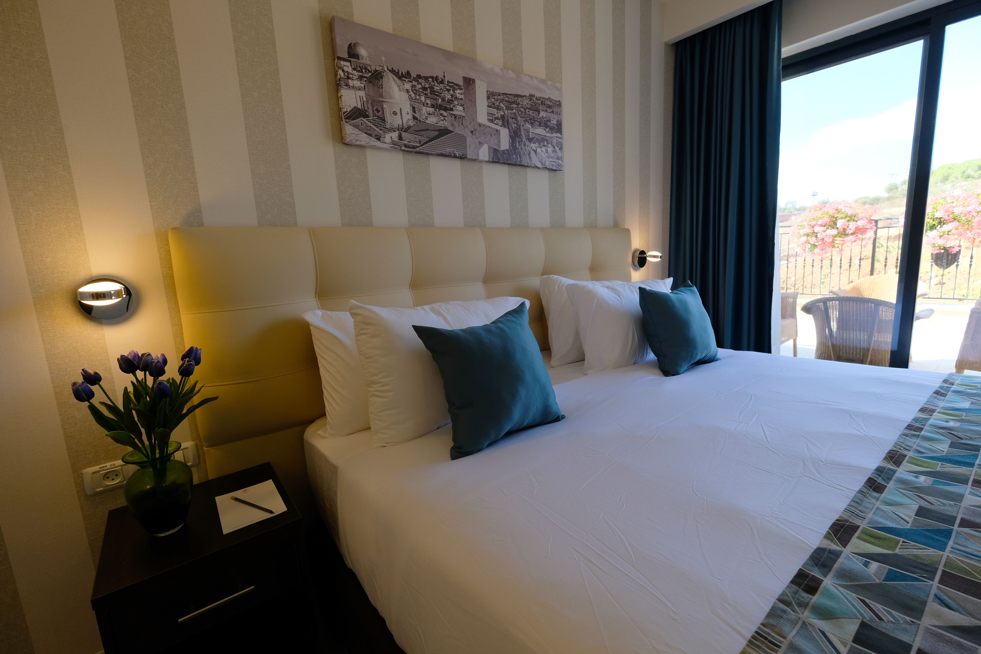 Une chambre d’hôtel avec un grand lit, une table de chevet ornée d’un vase de fleurs et une vue sur un balcon fleuri.