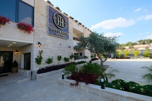 Una entrada de hotel con fachada de piedra, un cartel que dice "TANTUR HILLS HOTEL", y una zona ajardinada con plantas y un árbol.