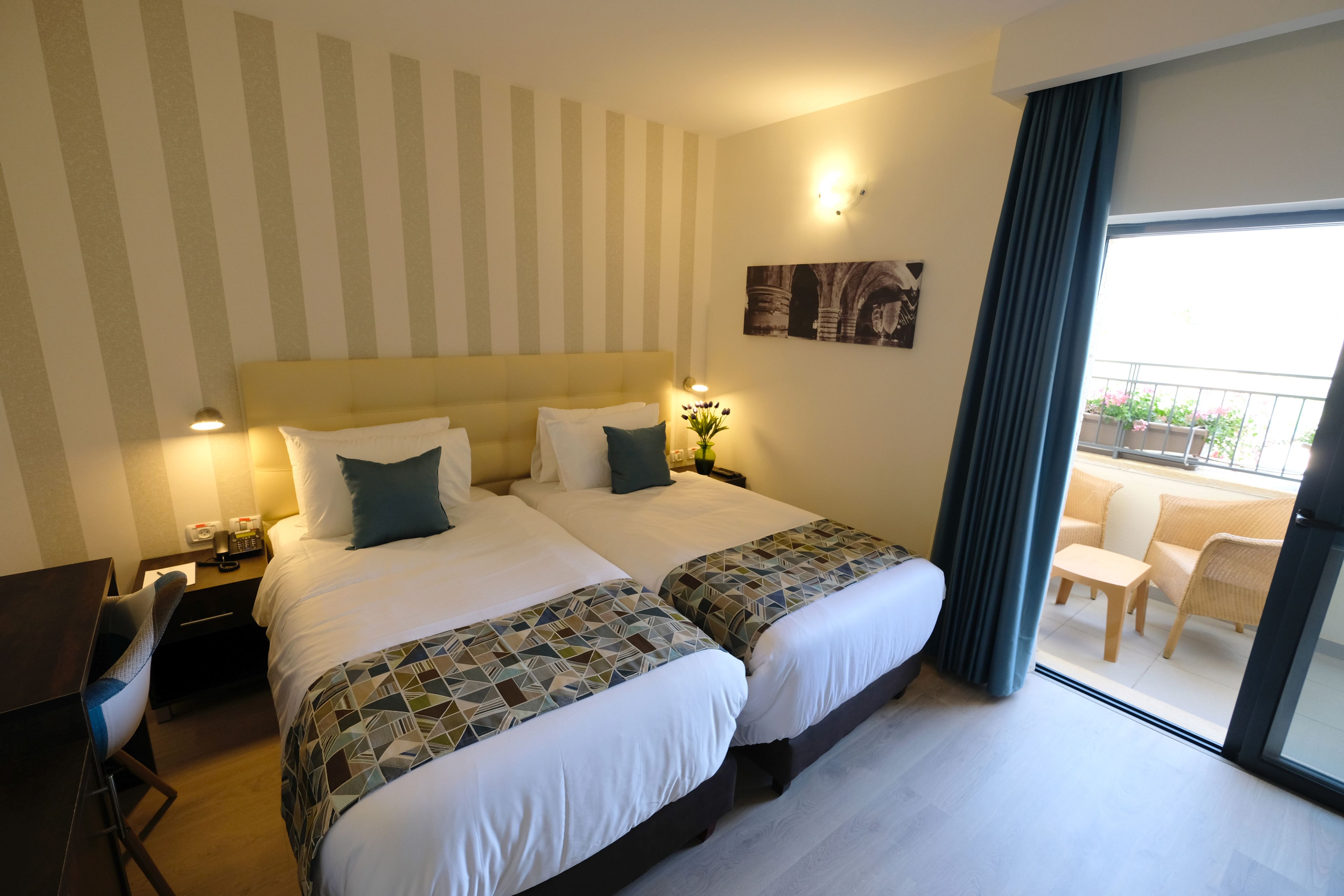 Habitación de hotel con dos camas, un escritorio y balcón con mobiliario exterior.