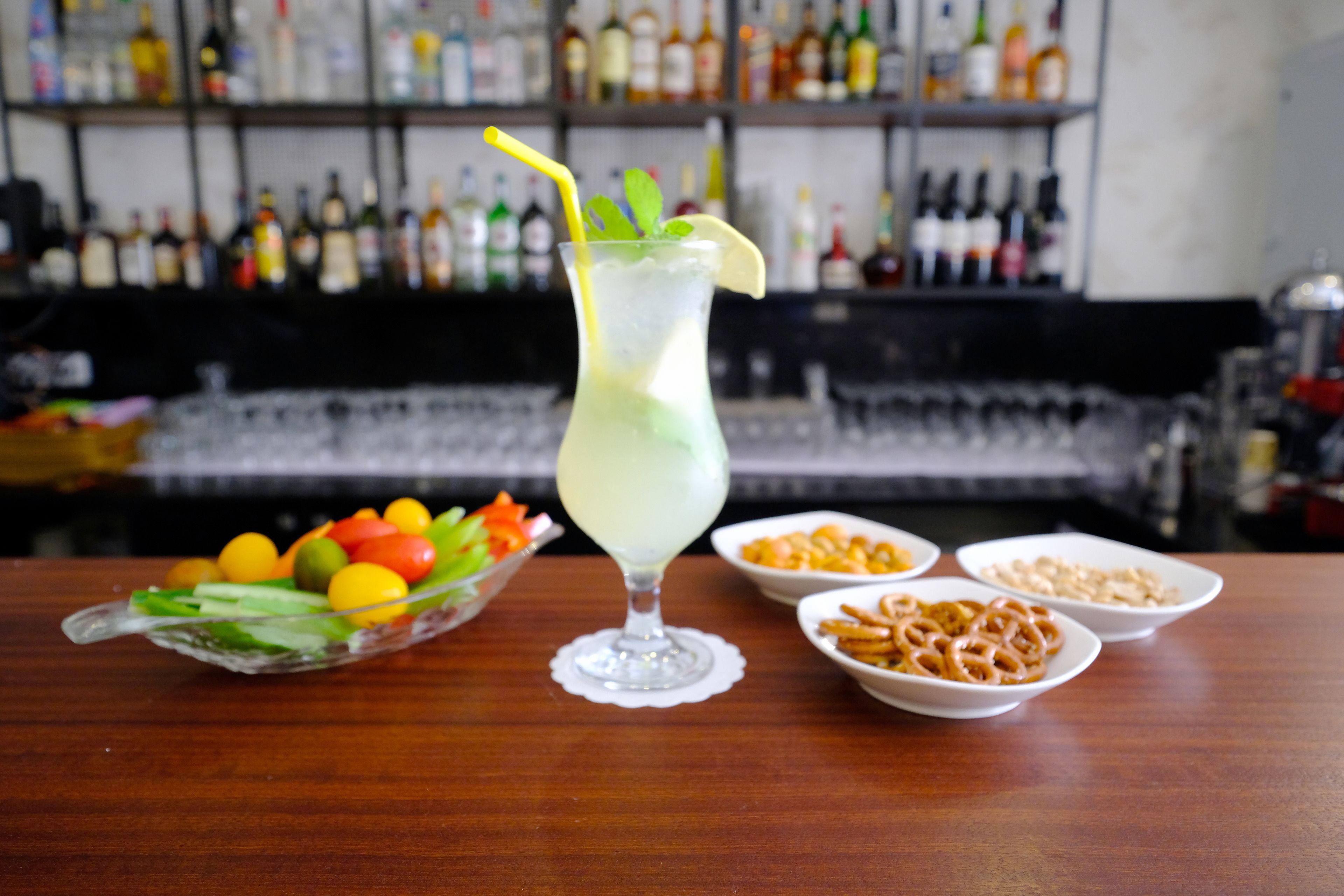 A bar setting with a tall glass drink, a bowl of fruit, and bowls of snacks on a wooden table.