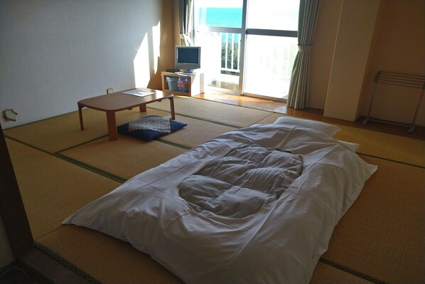 A room with tatami flooring, a bed with white bedding, a small table with a chair, a television on a stand, and a window with curtains.