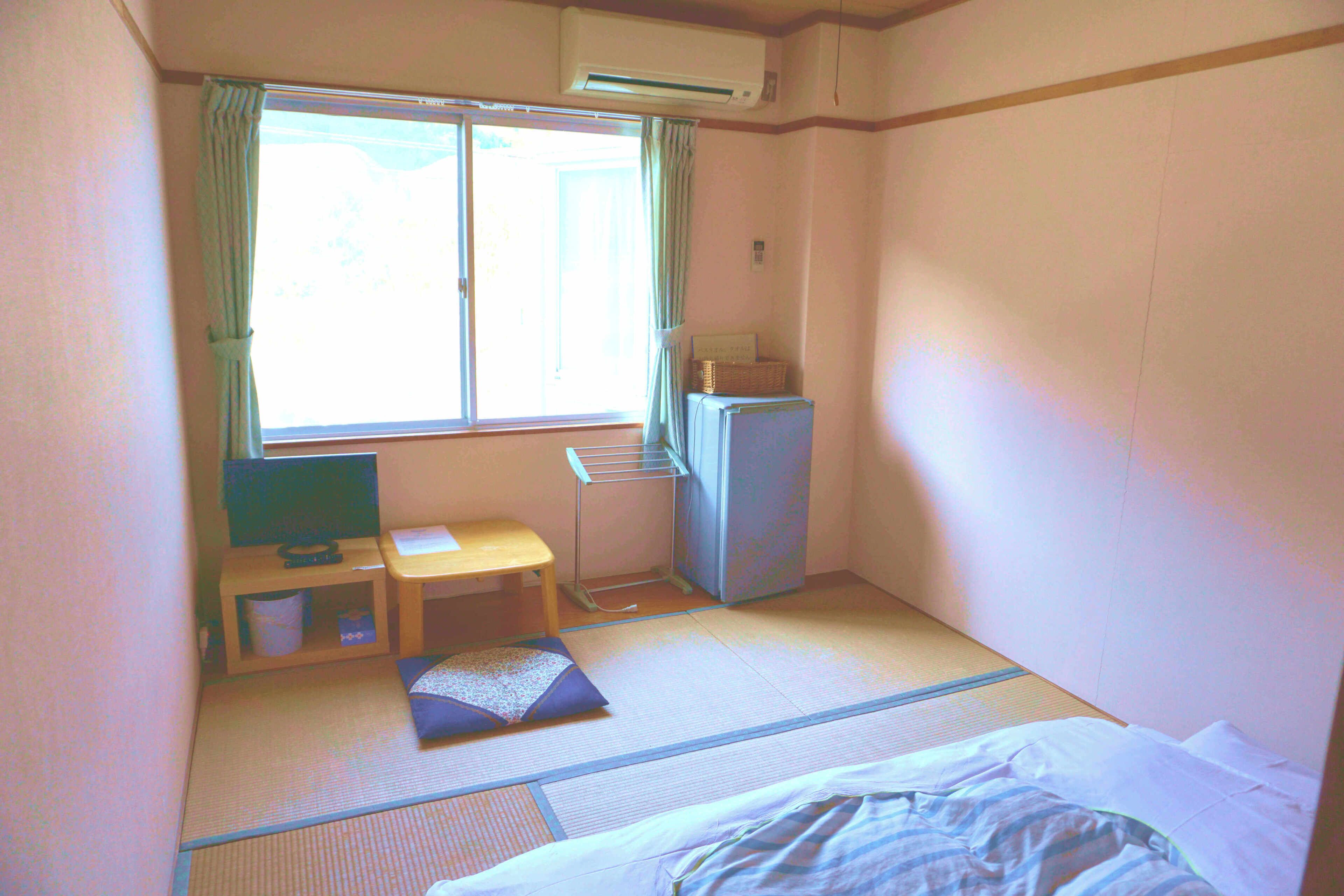 Quarto com piso de tatame, cama, televisão sobre uma mesinha, frigorífico e janela com cortinas.