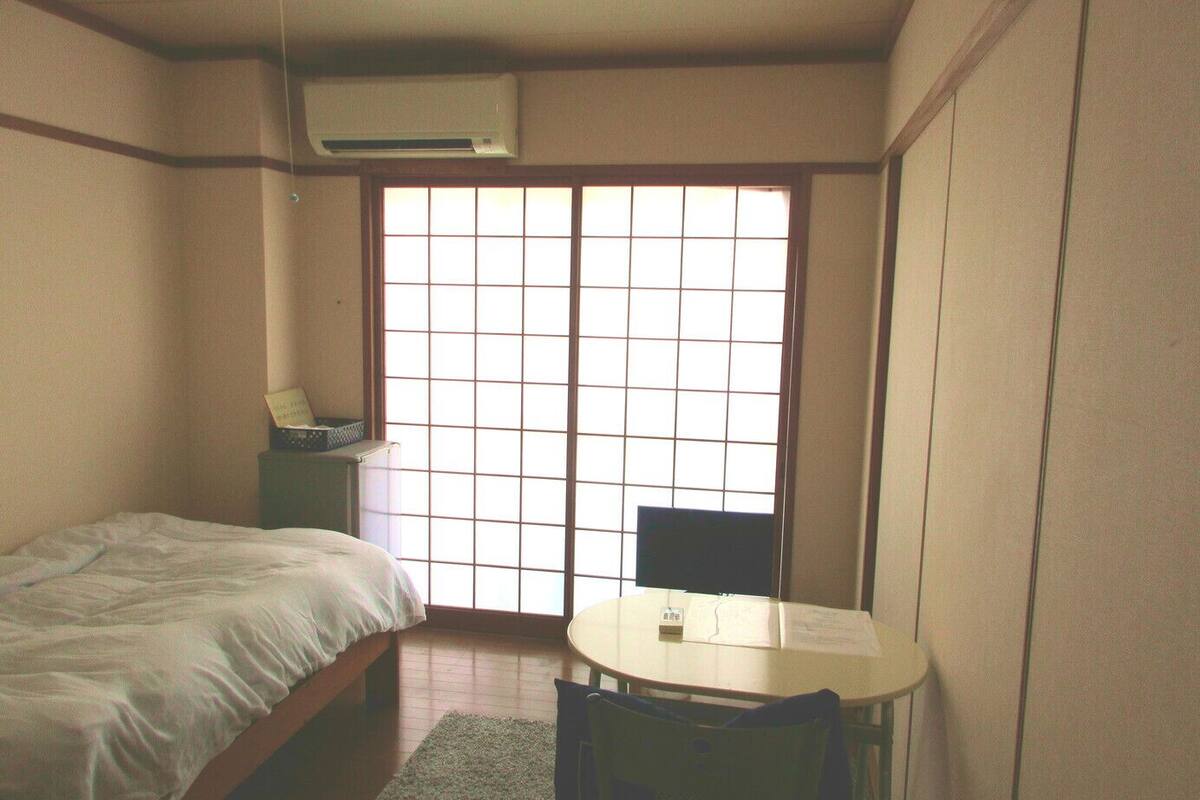A small room with a bed, a desk, and a chair. There is a window with a grid pattern.