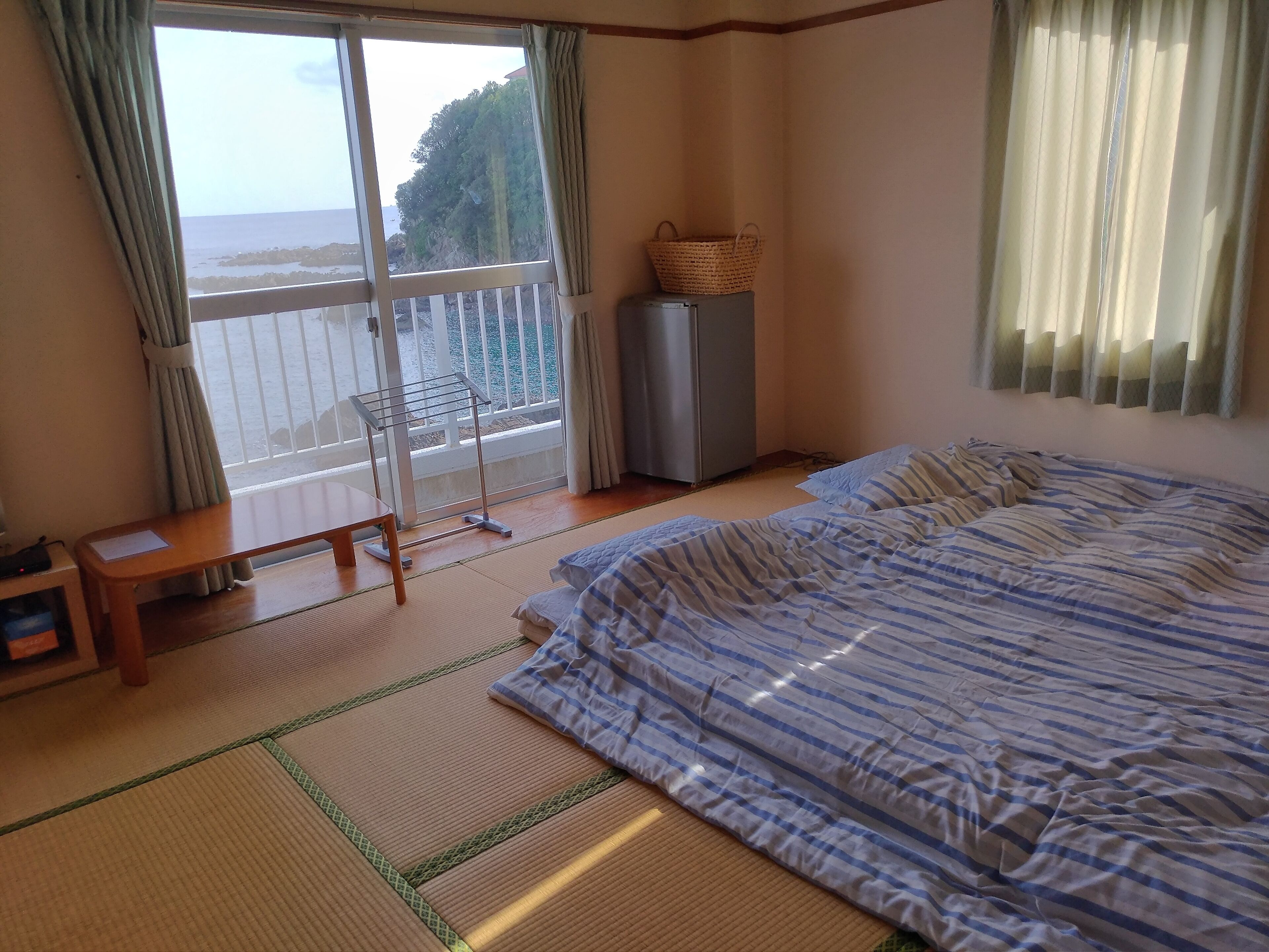 A room with a bed, a small table, a chair, a window with a view of the sea, and a balcony with a drying rack.