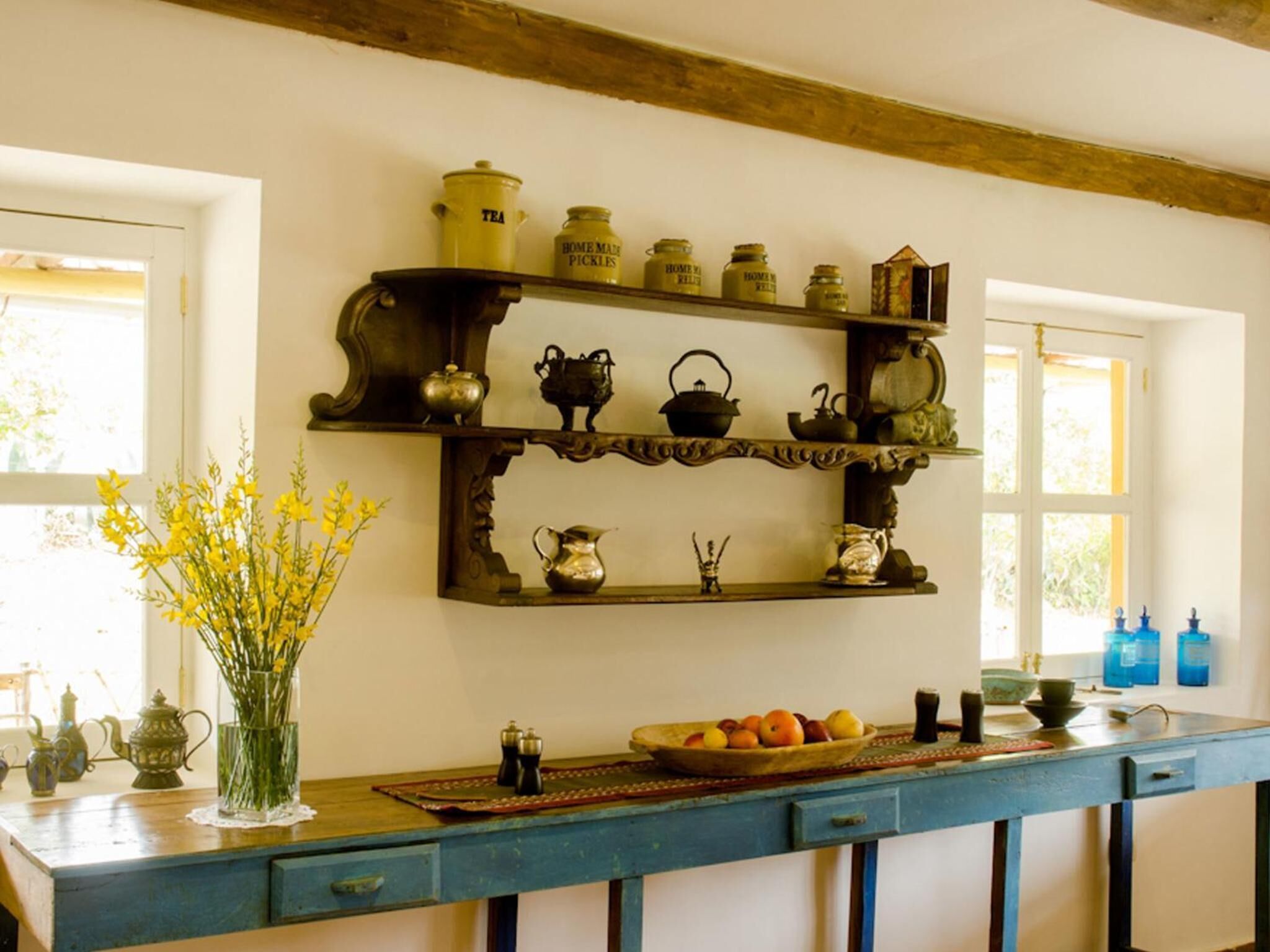 A kitchen with wooden shelves holding various kitchenware, a vase with yellow flowers, and a fruit bowl on the counter.