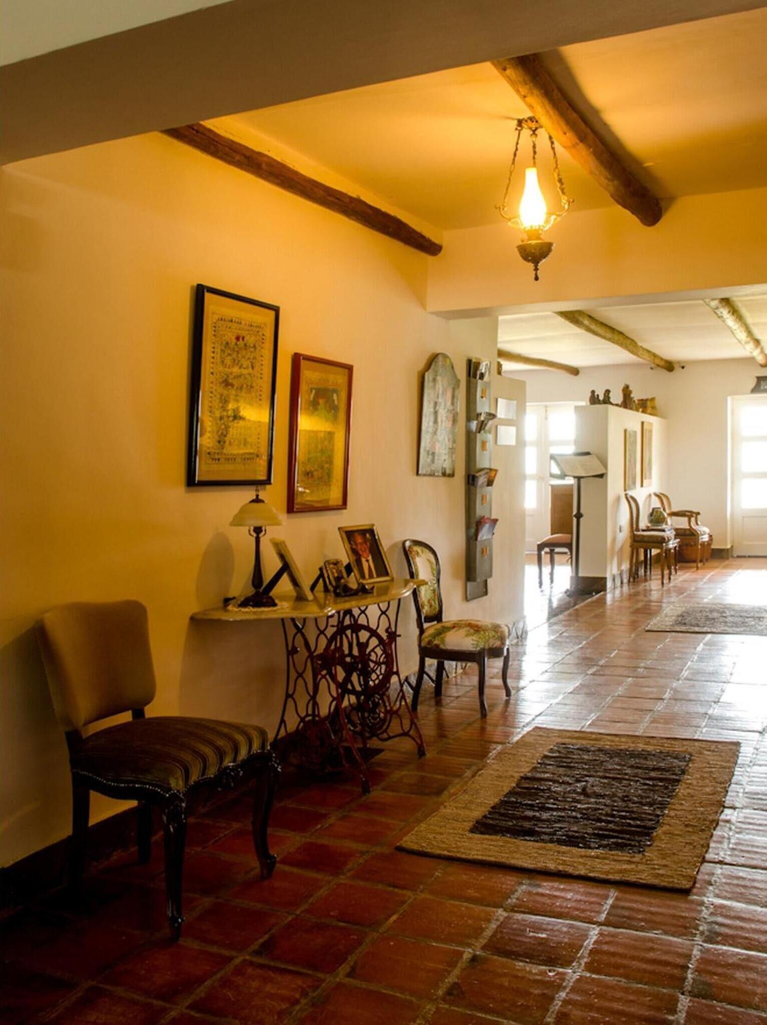 A spacious hallway with a tiled floor, a wooden chair, a table with a lamp, and framed artwork on the wall.