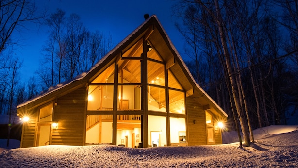 Una cabaña de madera con ventanas amplias, un balcón y terreno cubierto de nieve.