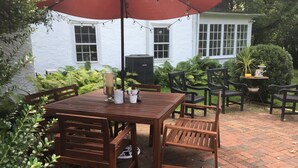 A patio with a wooden table and chairs, surrounded by lush greenery and a white building in the background. - Great location for college visits/move-in, parents weekend, Merion Golf, Phila! (Ardmore)