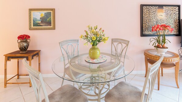 Comedor con mesa de vidrio rodeada de sillas blancas, un jarrón con flores amarillas y un cuadro en la pared.