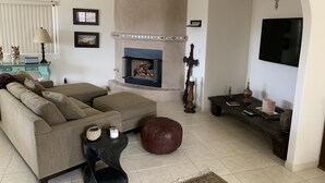A living room with a sofa, ottoman, fireplace, and a TV mounted on the wall. - Tranquil Desert Retreat . Short walk to hot springs & close to Joshua Tree! (Desert Hot Springs)