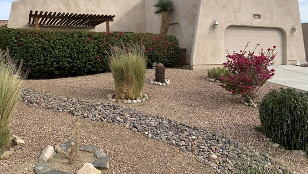 A building with a beige stucco exterior, a satellite dish, a wooden pergola, and a gravel driveway with various plants and a cactus. - Tranquil Desert Retreat . Short walk to hot springs & close to Joshua Tree! (Desert Hot Springs)