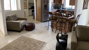 A modern living room with a sofa, ottoman, and a coffee table. There is a kitchen area with a refrigerator, stove, and bar stools. - Tranquil Desert Retreat . Short walk to hot springs & close to Joshua Tree! (Desert Hot Springs)