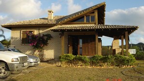 A two-story house with a tiled roof, a wooden porch, and a brick chimney. There are two cars parked in front. - Beautiful house in Praia do Rosa overlooking the lagoon. (Praia do Rosa)