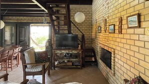 A cozy living room with a brick fireplace, wooden furniture, and a television. - Beautiful house in Praia do Rosa overlooking the lagoon. (Praia do Rosa)