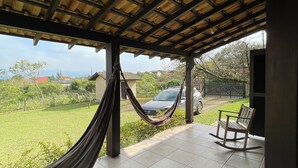 A covered patio with a hammock, a rocking chair, and a view of a green lawn and parked cars. - Beautiful house in Praia do Rosa overlooking the lagoon. (Praia do Rosa)