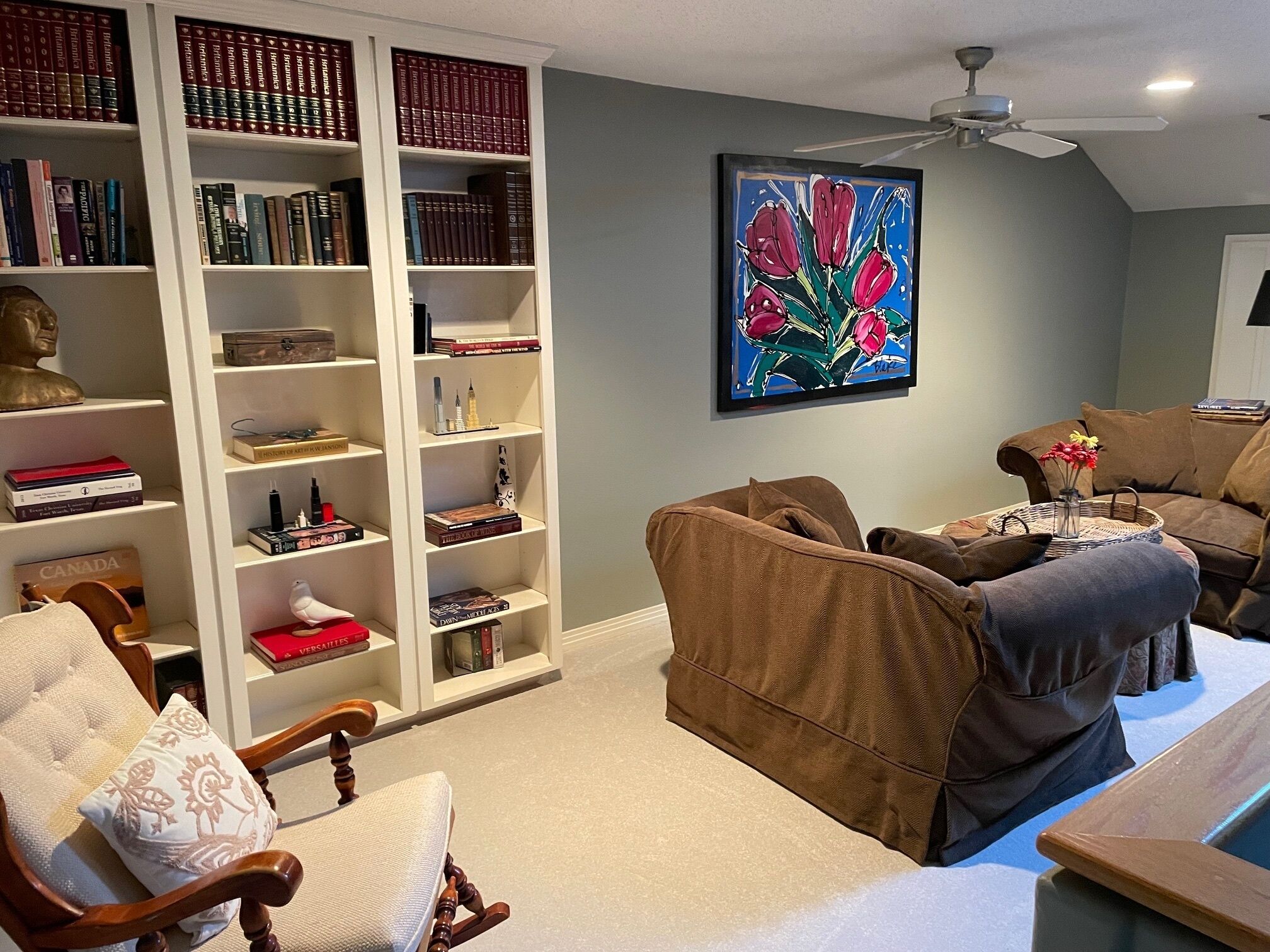 A living room with a bookshelf, a painting, and a ceiling fan.