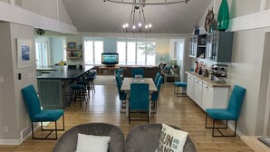 Uma sala ampla com área de estar central, cozinha com balcão e espaço de jantar com cadeiras azuis.