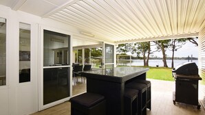 Une terrasse extérieure dotée d’un espace repas fermé par des baies vitrées, d’un barbecue et offrant une vue sur un lac.