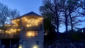 A two-story building with a balcony on the upper floor, a covered porch with string lights, and a dock leading to a body of water. - Lake Condo- Right on the Water with a Dock! Bring a boat or rent a boat! (Clear Lake)