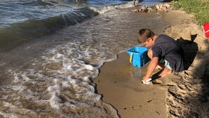 Escena de playa con un muelle de madera que se adentra en el agua, un niño jugando cerca de la orilla y un cubo rojo sobre la arena.