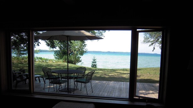 Habitación con un ventanal grande que da a una terraza con mesa y sillas, y vista a un cuerpo de agua.