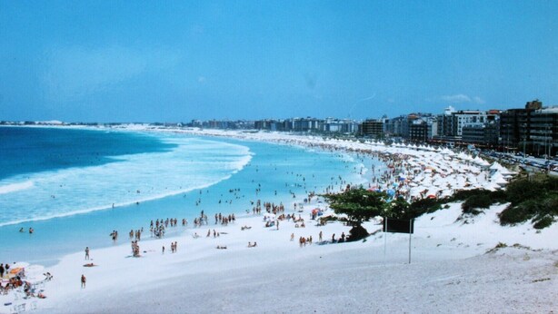 Una playa concurrida, un mar azul cristalino y edificios a lo largo de la costa.