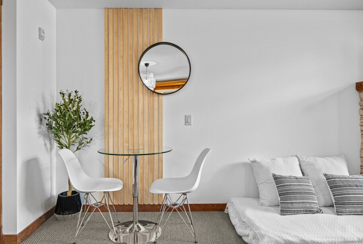 A modern room with a glass table and two white chairs, a bed with white and gray pillows, and a mirror on the wall.