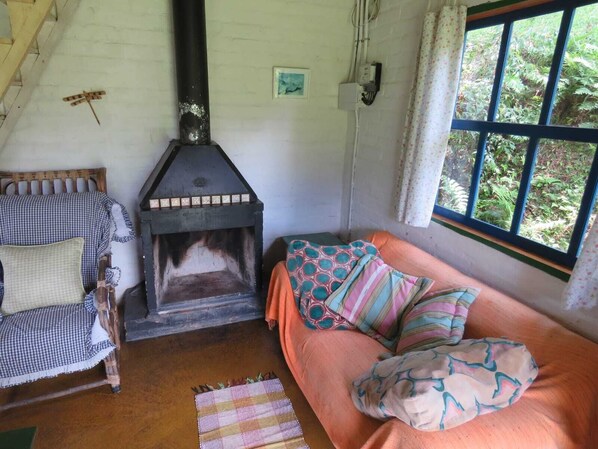 A cozy room with a fireplace, a wooden chair, a sofa with patterned cushions, and a window with a view of greenery.