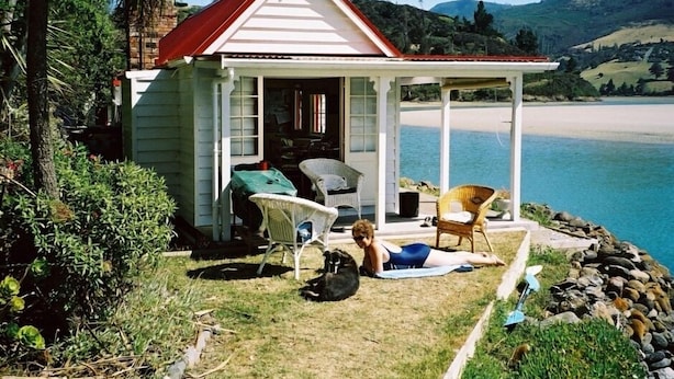 A small white house with a red roof, a porch with chairs, and a view of a beach and hills.