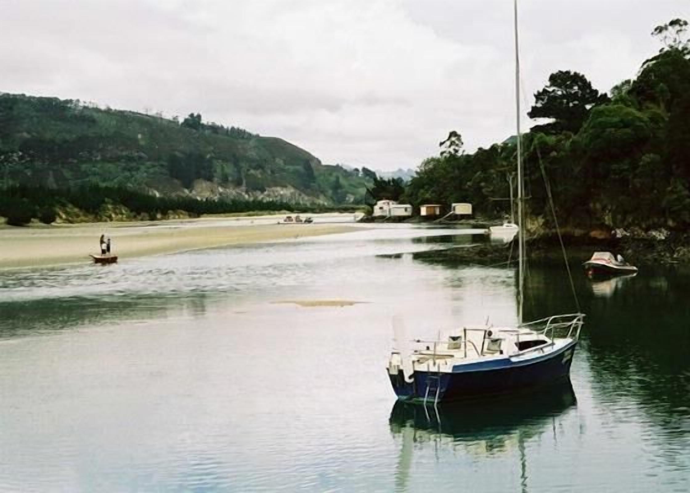 A calm river with a boat anchored near the shore, surrounded by greenery and a few buildings in the background.