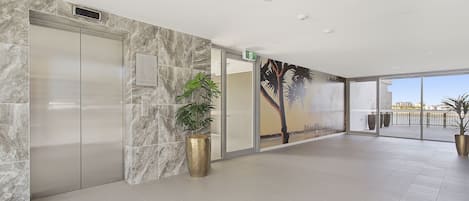 Un ascensor moderno, una planta en maceta grande y un mural de palmeras.